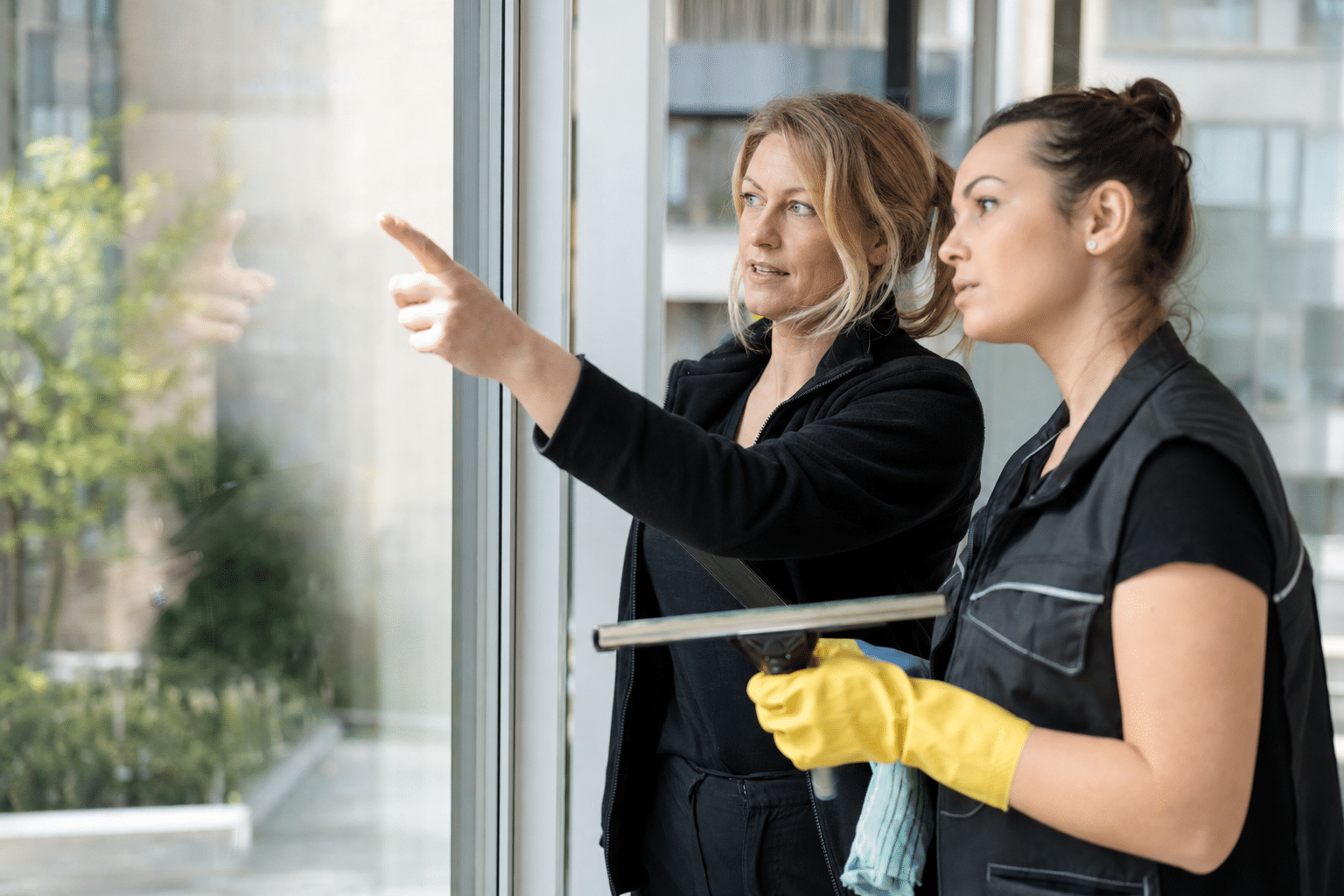 Objektleiterin prüft gemeinsam mit einer Reinigungskraft eine Glasfläche im Streiflicht